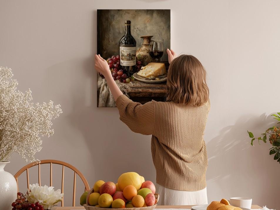 A woman hangs a framed painting of a still life of wine, cheese, and fruit on a wall in a cozy, homely setting.