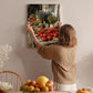 A woman hangs a framed painting of a fruit stand on a wall in a cozy kitchen setting.