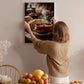 A woman hangs a framed photograph of a bowl of soup on a wall in a cozy kitchen setting.