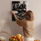 A woman hangs a framed photograph of a table with various dishes, including bowls, plates, and a vase, on a wall in a room with a dining table and chairs.