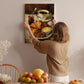 A woman hangs a painting of a breakfast scene on a wall, featuring a plate of croissants, fruit, and coffee.
