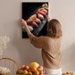 A woman hangs a framed photograph of sushi on a wall in a room with a dining table, chairs, and various fruits.