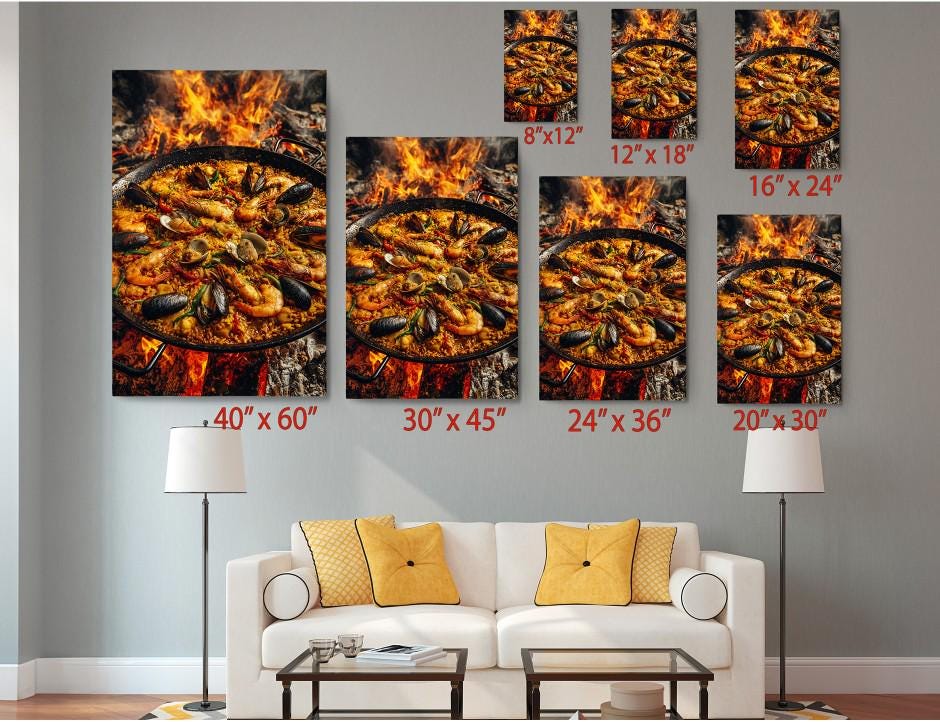 a living room with a white sofa and a coffee table. Above the sofa, there are several framed prints of the same image, depicting a large pan of seafood on a fire. The prints vary in size, ranging from 6 x 12" to 24 x 36". The prints are arranged in a grid-like pattern on the wall, creating a visually appealing display.