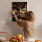 A woman hangs a framed photograph of a kitchen scene on a wall, surrounded by various fruits and flowers in a cozy, homely setting.