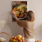 A woman hangs a framed photograph of a plate of food on a wall in a cozy kitchen setting.