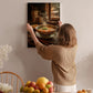 A woman hangs a framed photograph of a bowl of soup on a wall in a cozy kitchen setting.
