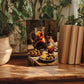 a still life painting of a table with a pitcher of wine, plates of olives, and glasses of wine, surrounded by potted plants and books.